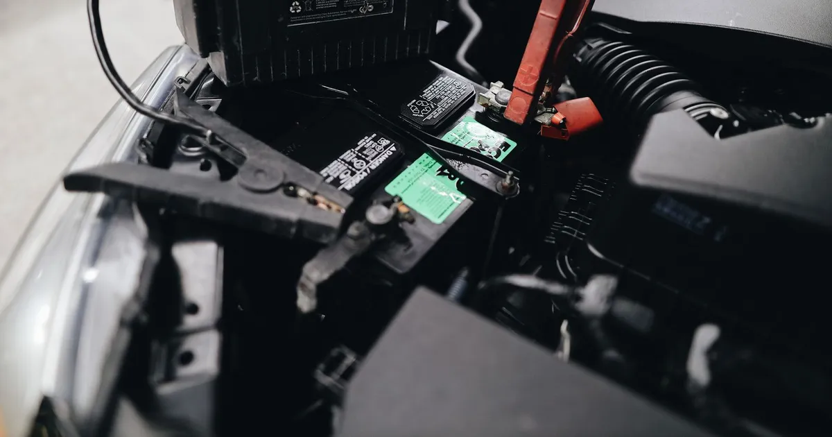 A 12V car battery in an engine bay with a jumper-cable clamp on the positive terminal