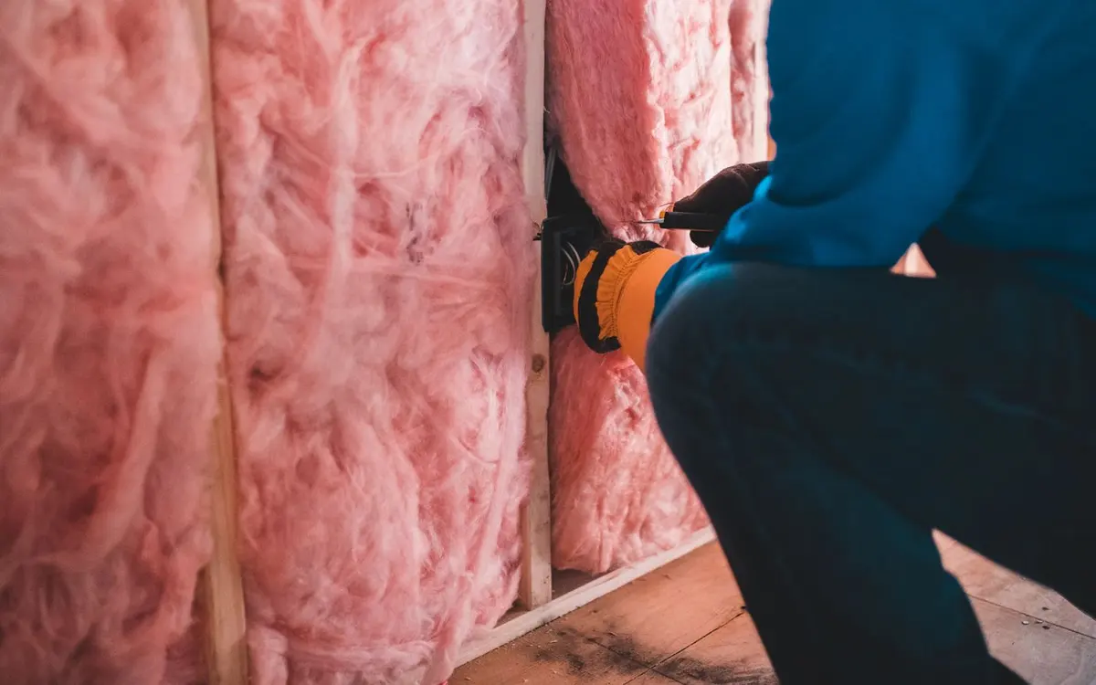 Fiberglass batt insulation packed between wood framing in a ceiling cavity, with a gloved installer working near an electrical box
